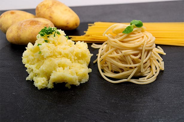Eine Portion gestampfte Kartoffeln neben einer Portion gekochter Nudeln. Im Hintergrund jeweils dahinter drei rohe Kartoffeln und ungekochte Spaghetti.