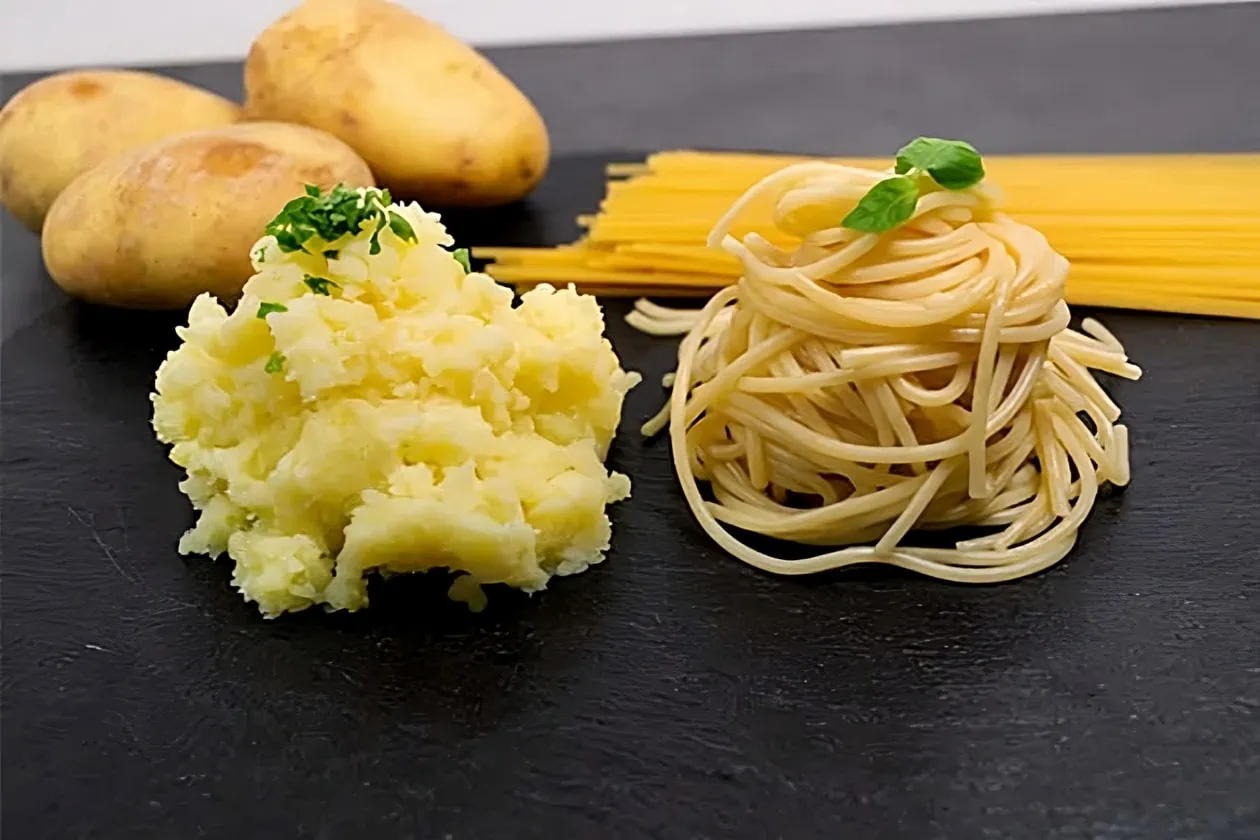 Eine Portion gestampfte Kartoffeln neben einer Portion gekochter Nudeln. Im Hintergrund jeweils dahinter drei rohe Kartoffeln und ungekochte Spaghetti.